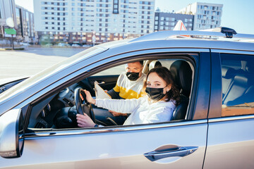 Brunette middle-eastern woman in face mask learning parking at driving school while COVID-19 pandemic, holding hands on automobile steering wheel, sitting by male driving instructor, side view