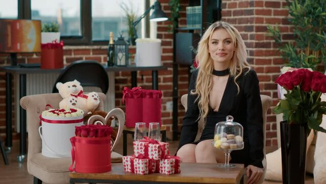 Gorgeous blonde in video call waving hello at loved one on valentine day happy with surprise gifts in livin room. Beautiful woman smiling saying hello at camera enjoying gift boxes and red roses.