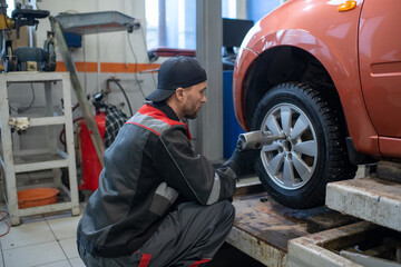 Young repairman in workwear using electric handtool while sitting on squats and changing wheel of car in garage