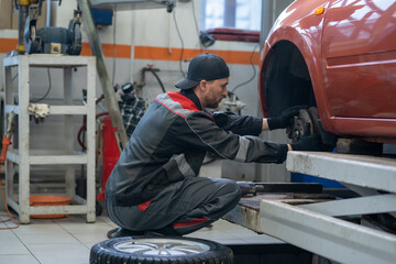 Contemporary repairman or technician in workwear changing wheel of car while sitting on squats in front of automobile