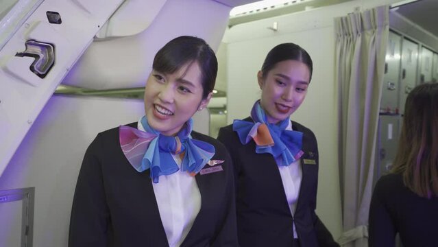 Two young beautiful Asian female flight attendants in suit uniform and scarfs, standing at the airplane entrance, smiled friendly checking passenger's boarding pass and welcoming them to the flight.