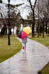 A cute little girl in a blue cape, pink boots and a pink hat runs through puddles and has a fun.The girl has a rainbow umbrella in her hands. Happy childhood. Early spring. Emotions.