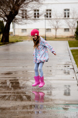 A cute little girl in a blue cape, pink boots and a pink hat runs through puddles and has a fun....