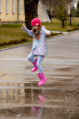 Fototapeta premium A cute little girl in a blue cape, pink boots and a pink hat runs through puddles and has a fun. Happy childhood. Early spring. Emotions.