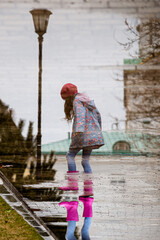 A cute little girl in a blue cape, pink boots and a pink hat runs through puddles and has a fun. Happy childhood. Early spring. Emotions.