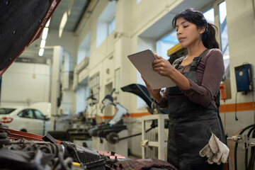 Young Hispanic woman in overalls looking at screen of digital tablet in her hands while repairing...