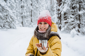 Fototapeta na wymiar A young girl smiles in the winter forest, a mug of coffee in her hand. The red cap is covered with snowflakes, the weather is cold. A woman in a yellow jacket walks through a snowy forest.