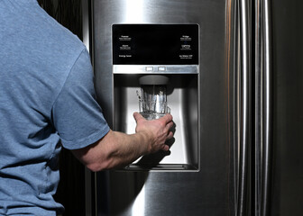 Male right hand filling glass with water splashing out of dispenser of home fridge