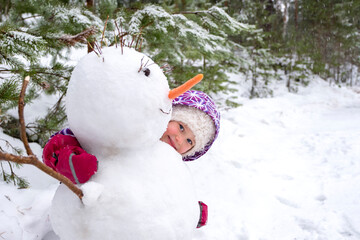 A little Caucasian girl 5 years old looks out from behind a snowman and smiles. Winter...