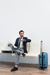 man with newspaper looking at wristwatch while sitting in departure lounge near suitcase.