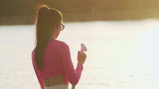 Back view of young woman with brown hair practising in sup boarding on city lake. Attractive female in sport clothes enjoying evening workout on fresh air. 
