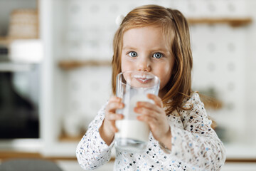 Cute little girl drinking fresh milk at home and looking at camera