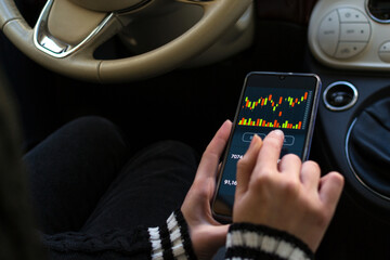 Young woman sitting in her car reviewing and investing in the stock market with technical indicator tools on his smartphone. Trading concept.