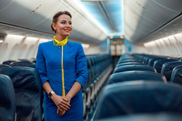 Cheerful woman stewardess in airline air hostess uniform standing in aisle of airplane passenger...