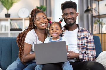 Portrait of beautiful and happy family in casual wear sitting together on comfy couch and using wireless laptop. African american parents spending time with their cute little daughter.