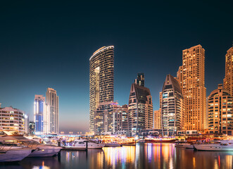 Fototapeta na wymiar Dubai Marina Port, UAE, United Arab Emirates. Jetty With Many Moored Yachts, Sightseeing Boat In Evening Night Illuminations. Night View Of Dubai Marina Skyline.