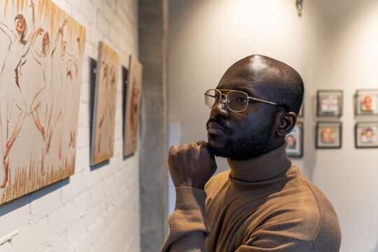 African pensive man in eyeglasses examining the artwork of famous artists standing in art gallery