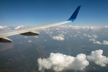 View of jet airplane wing from inside flying over white puffy clouds in blue sky. Travel and air transportation concept.
