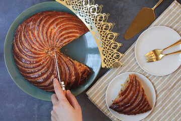 Top view of French galette des rois, hand holding knife cutting it in a piece with crown, white plates and cutlery. It's made with puff pastry and creamy almond filling roll in circles shape. 