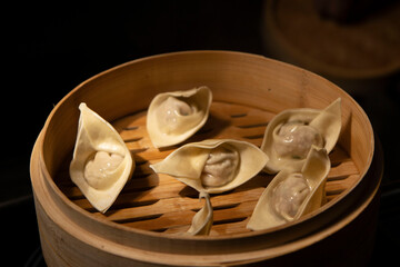 Fresh wonton dumplings with meat are presented close-up in a bamboo steamer against the dark background of the restaurant. Chinese cuisine. Asian steam food dim sum, stuffed dumplings.