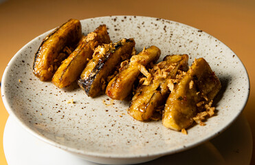 Caramelized fried bananas presented on a round gray plate close up on a brown background. Crispy banana pieces fried in butter and sugar. Restaurant serving food, sweet Thai dessert. Asian cuisine.