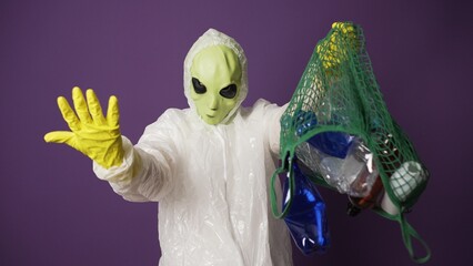Humanoid with green skin wearing sanitary suit and rubber gloves throws empty plastic bottles out of mesh bag against violet wall closeup