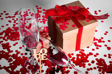 close up of two gift boxes with red ribbon, red confetti, two champagne glasses on a white background