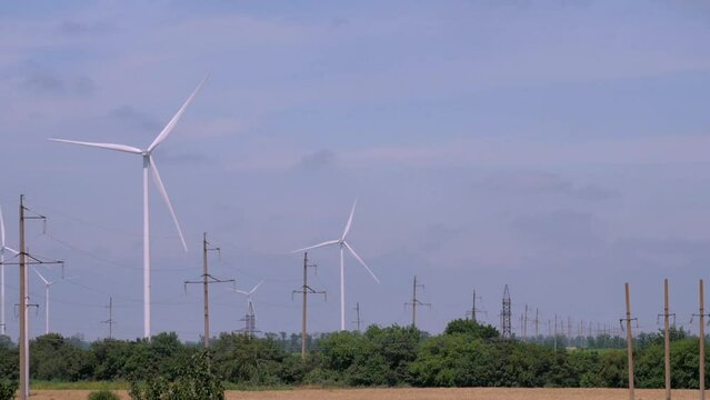 Wind energy production at rural station. Powerful propellers rotate in field with electric power lines near lush treen trees on cloudy day