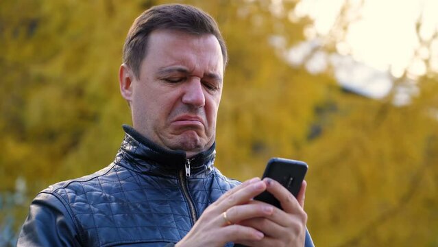 Sad millennial guy looks at phone screen in surprise and stands outside at autumn. Disgusted and overwhelmed young man stares at screen of smart phone, needs to pay debts immediately