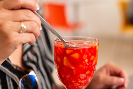 Hand of an adult woman using a long spoon to eat a tropical red fruit drink. Colombian drink. Salpicon