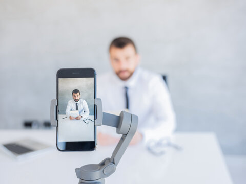 Young businessman with beard sitting in office recording himself on the phone which is in the focus while he is in the background blurred.