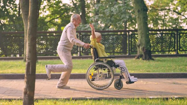 Active senior man running pushing wheelchair with happy wife in park, happiness
