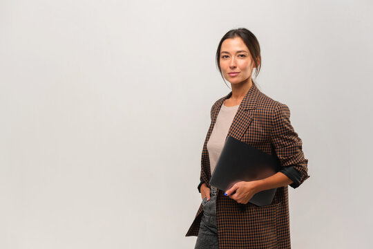 Stylish confident asian female standing with laptop in her hands and looking at camera at gray background. Portrait of sophisticated woman advertising products and services