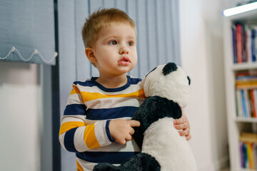 Waist up portrait of one small caucasian boy two years old holding panda toy while standing alone...