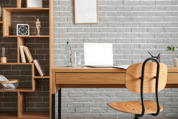 Comfortable workplace with modern laptop, bottle of water and glass near grey brick wall