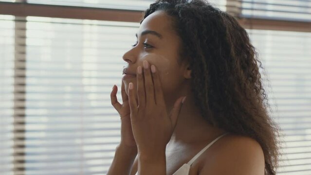 Skin care. Young african american woman applying nourishing cream on her cheeks, smiling to herself at bathroom, zoom in