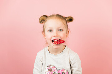 Funny little blonde girl smiling and playing with red heart shape lollipop caramel on pink background. Valentines Day or Mothers Day concept