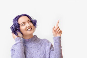 Beautiful young woman with violet hair and fluffy headphones pointing at something on white...