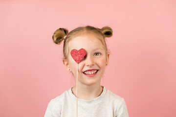 Funny little blonde girl smiling and playing with red heart toy on pink background. Valentines Day or Mothers Day concept