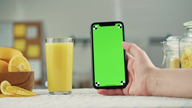 Woman pouring orange juice and holding smartphone with chroma key, fresh juice on kitchen table, vitamin c, cooking drink made of citrus fruits. Healthy eating habits in morning.