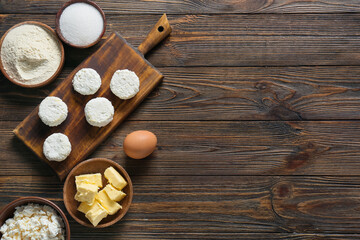 Board with uncooked cottage cheese pancakes and ingredients on wooden background