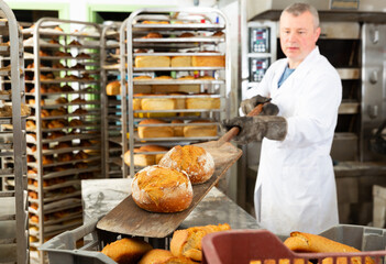 Portrait of focused man engaged in breadmaking, taking out ready loaves from oven in bakeshop..