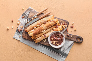 Wooden board of tasty thin pancakes with chocolate paste and nuts on beige background
