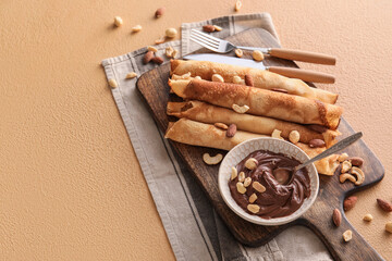Wooden board of tasty thin pancakes with chocolate paste and nuts on beige background