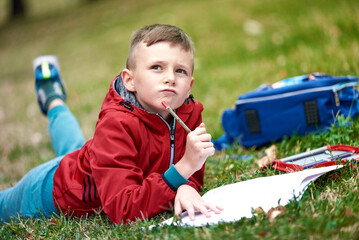 Cute young boy writing his homework in a park, lying on the grass, happy to learn and enjoying a nice sunny day in the park