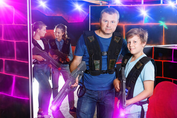 Portrait of glad cheerful positive teen boy and his father standing indoors with laser guns ready for lasertag game.