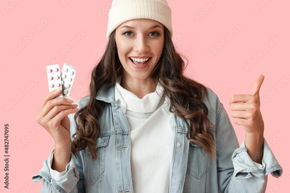 Wall mural Young happy woman showing thumb up and holding blisters with pills on color background