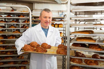 Skilled successful baker arranging trays with freshly baked bakery products on trolley..