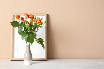 Vase with beautiful orange roses and blank photo frame on table against beige wall