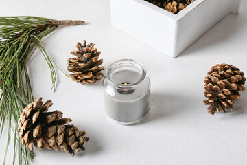 Glass holder with burning candle and pine cones on light background